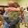 A military homecoming event taking place in what appears to be a community hall or gymnasium. A soldier in camouflage uniform is embracing a young girl who has her arms and legs wrapped around him in an emotional reunion. The scene is being documented by a videographer, and numerous family members and bystanders are present, many visibly emotional.