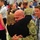 A military homecoming or deployment ceremony is taking place in what appears to be a large indoor venue decorated with American flags. A bald soldier in camouflage uniform is embracing a person in a black top, while family members and other reuniting couples surround them. The emotional scene captures multiple reunions happening simultaneously, with media personnel visible recording the event.