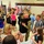 A military homecoming ceremony taking place in what appears to be a community center or armory gymnasium. Multiple soldiers in camouflage uniforms are being reunited with family members, with emotional embraces visible throughout the room. American flags are prominently displayed along the walls, and media personnel with cameras are documenting the event.