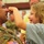 A U.S. Army soldier in MultiCam/OCP camouflage uniform is sharing a joyful moment with a young girl, who is playfully touching his nose. Both are smiling and laughing, suggesting a warm reunion or family gathering. The background shows additional people in a large indoor venue, consistent with a military homecoming or send-off ceremony.