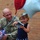 A U.S. Army soldier named Knippel, wearing OCP (Operational Camouflage Pattern) uniform, crouches beside a young girl who is holding balloons and a small gift. The scene appears to be a military homecoming or welcome event, suggested by the patriotic red, white, and blue decorations including star-shaped balloons and confetti streamers. Both individuals are smiling, conveying a joyful reunion atmosphere.