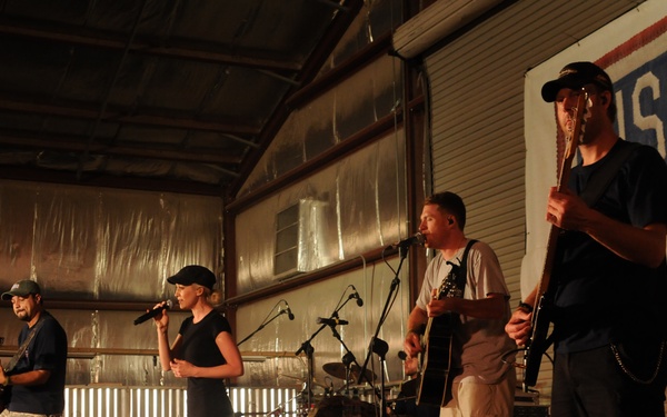 A live band performance is taking place inside a metal hangar or warehouse-style building at what appears to be a USO show. Four performers are visible on stage: two guitarists, a female vocalist, and an acoustic guitarist, with a drum kit visible in the background. A large USO banner is prominently displayed on the right side of the venue.