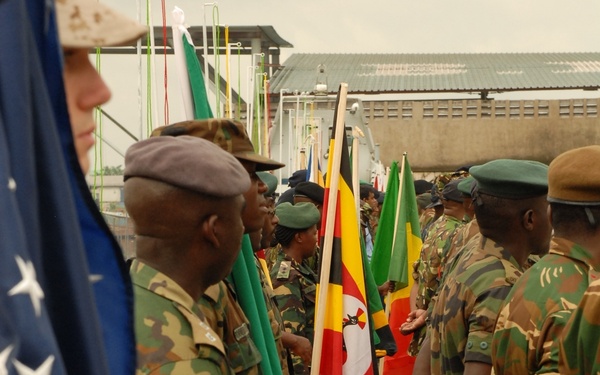 A multinational military ceremony or parade featuring soldiers from multiple nations standing in formation while carrying their respective national flags. The Uganda flag is prominently visible in the center, alongside the United States flag on the left edge. Soldiers are wearing various camouflage uniforms and military headgear, suggesting a joint military exercise or official ceremony.