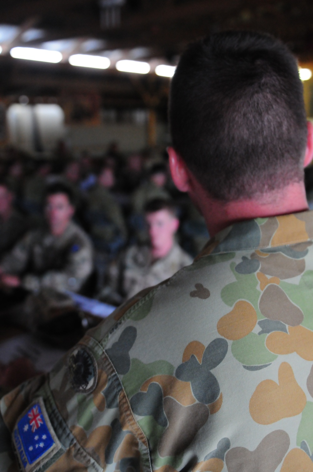 A military briefing or assembly is taking place inside what appears to be a hangar or large indoor facility. The foreground shows the back of an Australian Defence Force (ADF) soldier wearing distinctive Australian Disruptive Pattern (AUSCAM) camouflage with a visible Australian flag patch on the sleeve. In the background, a group of seated military personnel are visible, suggesting a formal address or briefing is in progress.