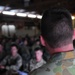 A military briefing or assembly is taking place inside what appears to be a hangar or large indoor facility. The foreground shows the back of an Australian Defence Force (ADF) soldier wearing distinctive Australian Disruptive Pattern (AUSCAM) camouflage with a visible Australian flag patch on the sleeve. In the background, a group of seated military personnel are visible, suggesting a formal address or briefing is in progress.