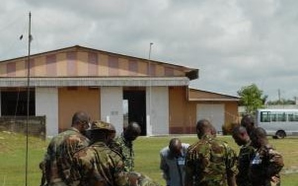 A group of military personnel in camouflage uniforms are gathered on a grass field, appearing to conduct a training exercise or equipment assembly. At least one civilian in blue clothing is present among the uniformed soldiers. In the foreground, a metal pole or antenna stake is visible on the ground along with wire or cable materials.