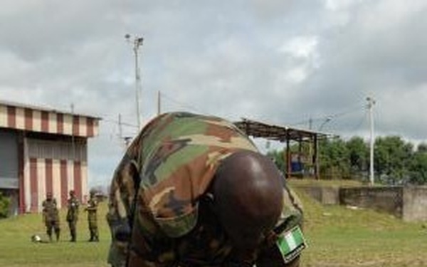 A soldier wearing camouflage uniform with what appears to be a Nigerian flag patch is bent over, using a sledgehammer or mallet to drive a stake or pipe into the ground. The activity takes place on a grassy field, with other uniformed personnel visible in the background. A striped building and utility poles are visible in the background setting.