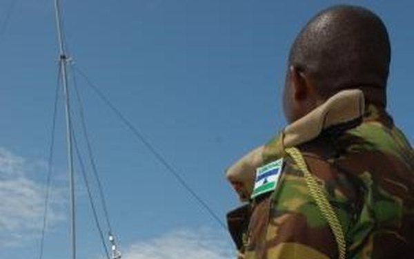 A military training or field exercise is taking place on an open grassy area. The foreground soldier wears a camouflage uniform with a shoulder patch appearing to be the flag of Sierra Leone, and is holding guide ropes connected to a tall antenna or mast being erected. Multiple soldiers in various uniforms are visible in the background assisting with the operation.