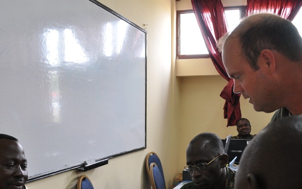 A group of military personnel from different nations are gathered in a conference or training room setting. A Caucasian officer in an olive green flight suit is leaning over a table engaging with African military officers, one of whom wears a uniform patch consistent with the Togolese Armed Forces. The setting suggests a multinational military cooperation meeting, briefing, or training exercise.