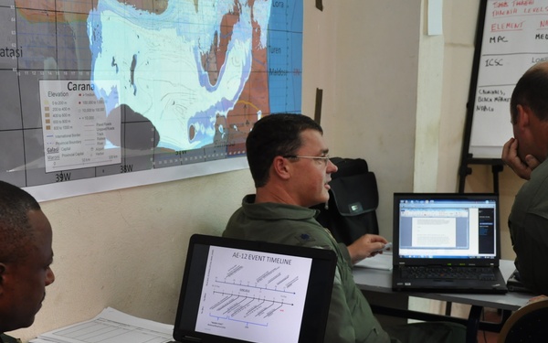 Three military personnel in uniform are gathered around a table in what appears to be a briefing or operations room. They are working with laptops, documents, and maps during what seems to be a planning or training exercise. A topographic map labeled 'Carana' is visible on the wall behind them.
