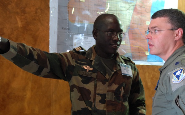 Two military officers are engaged in a briefing or planning session, with one officer in camouflage uniform pointing toward a map on the wall while the other in an olive flight suit listens attentively. A name tag reading 'DIAGNE' is visible on the camouflage-uniformed officer, and the flight-suited officer wears a unit patch on his left shoulder. A third individual is seated at a desk in the foreground, taking notes.