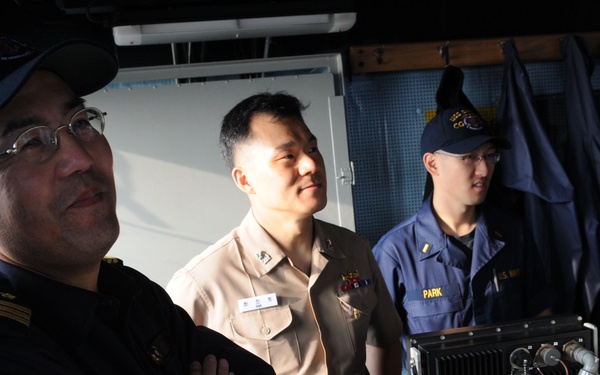 The image shows four military personnel inside what appears to be a naval vessel's bridge or operations room. At least two different uniforms are visible: a dark navy uniform worn by the person on the left, and U.S. Navy working uniforms (NWUs) worn by others. One person on the right is wearing a headset and appears to be operating equipment.