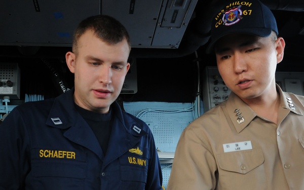 Two naval officers — one U.S. Navy and one appearing to be from the Republic of Korea Navy — are reviewing a nautical chart or map together on what appears to be a ship's bridge. The U.S. sailor is identified by his uniform nameplate as 'SCHAEFER' and wears a dark navy working uniform, while the other officer named 'LEE' wears a tan/khaki uniform and a cap reading 'USS SHILOH CG-67'. This appears to be a joint naval cooperation or exchange activity.