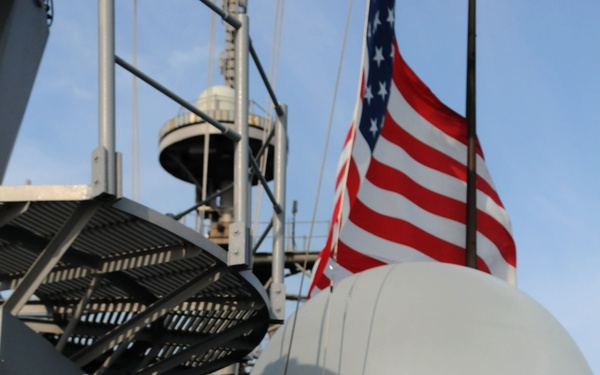 The image shows military personnel on the deck of a U.S. Navy warship, identifiable by the American flag flying prominently in the background. Personnel are wearing caps with ship designations including 'USS Shiloh CG-67' and what appears to be 'JDS Kirishima', suggesting a joint U.S.-Japanese naval exercise or visit. A large radar dome and various ship superstructure elements are visible in the background.