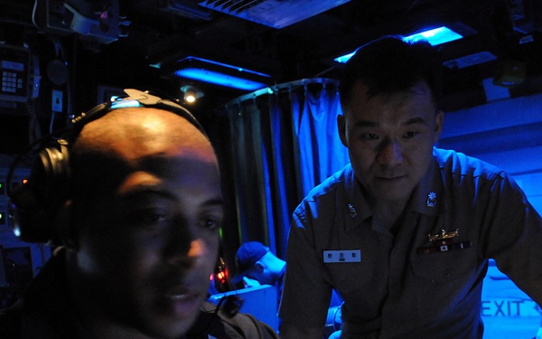 Two military personnel are working at a console station in what appears to be a naval operations center or combat information center (CIC) aboard a ship. The environment is dimly lit with blue ambient lighting, typical of a shipboard tactical operations space. One person in the foreground wears headphones and a dark uniform, while a second person in a khaki uniform leans over the console.