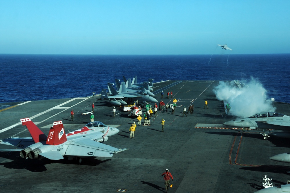 A busy U.S. Navy aircraft carrier flight deck showing active flight operations in progress. Multiple F/A-18 Hornet/Super Hornet jets are staged on deck, with at least one aircraft visibly launching via catapult, evidenced by steam/vapor from the catapult system. Deck crew in color-coded jerseys are performing various flight deck roles in open ocean conditions.