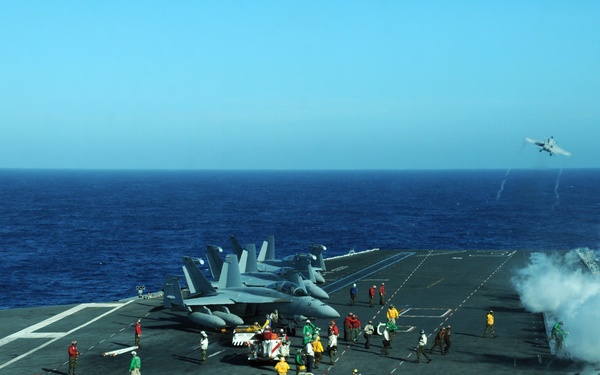 A busy U.S. Navy aircraft carrier flight deck showing active flight operations in progress. Multiple F/A-18 Hornet/Super Hornet jets are staged on deck, with at least one aircraft visibly launching via catapult, evidenced by steam/vapor from the catapult system. Deck crew in color-coded jerseys are performing various flight deck roles in open ocean conditions.