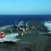 A busy U.S. Navy aircraft carrier flight deck showing active flight operations in progress. Multiple F/A-18 Hornet/Super Hornet jets are staged on deck, with at least one aircraft visibly launching via catapult, evidenced by steam/vapor from the catapult system. Deck crew in color-coded jerseys are performing various flight deck roles in open ocean conditions.