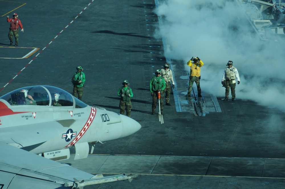 A U.S. Navy carrier-based jet aircraft, marked with the number '100' and featuring a red checkered nose design, is positioned on an aircraft carrier flight deck preparing for launch. Multiple flight deck crew members in color-coded jerseys (green, yellow, white, and red) are visible performing pre-launch duties. Steam or exhaust vapor is billowing from the catapult track area, indicating an imminent or recent catapult launch sequence.