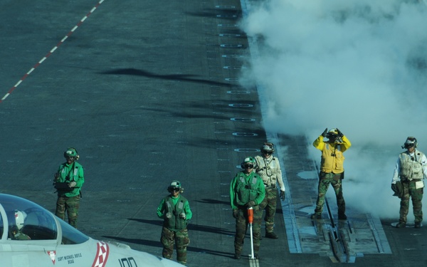 A U.S. Navy carrier-based jet aircraft, marked with the number '100' and featuring a red checkered nose design, is positioned on an aircraft carrier flight deck preparing for launch. Multiple flight deck crew members in color-coded jerseys (green, yellow, white, and red) are visible performing pre-launch duties. Steam or exhaust vapor is billowing from the catapult track area, indicating an imminent or recent catapult launch sequence.