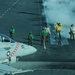 A U.S. Navy carrier-based jet aircraft, marked with the number '100' and featuring a red checkered nose design, is positioned on an aircraft carrier flight deck preparing for launch. Multiple flight deck crew members in color-coded jerseys (green, yellow, white, and red) are visible performing pre-launch duties. Steam or exhaust vapor is billowing from the catapult track area, indicating an imminent or recent catapult launch sequence.