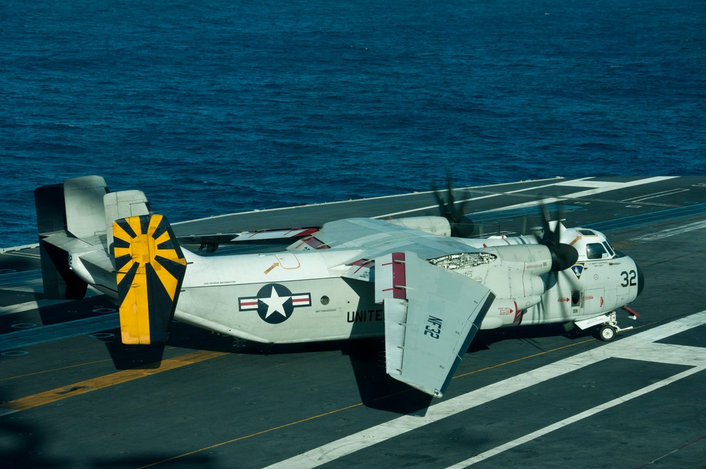 A U.S. Navy Grumman C-2 Greyhound carrier onboard delivery (COD) aircraft, marked with the number 32, is parked on the flight deck of an aircraft carrier at sea. The aircraft features folded wings, a distinctive yellow and black sunburst tail marking, and U.S. military insignia. The ocean is visible in the background, indicating the carrier is underway.