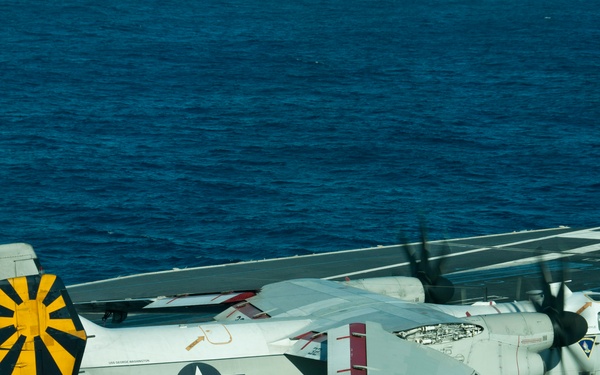 A U.S. Navy Grumman C-2 Greyhound carrier onboard delivery (COD) aircraft, marked with the number 32, is parked on the flight deck of an aircraft carrier at sea. The aircraft features folded wings, a distinctive yellow and black sunburst tail marking, and U.S. military insignia. The ocean is visible in the background, indicating the carrier is underway.