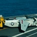 A U.S. Navy Grumman C-2 Greyhound carrier onboard delivery (COD) aircraft, marked with the number 32, is parked on the flight deck of an aircraft carrier at sea. The aircraft features folded wings, a distinctive yellow and black sunburst tail marking, and U.S. military insignia. The ocean is visible in the background, indicating the carrier is underway.