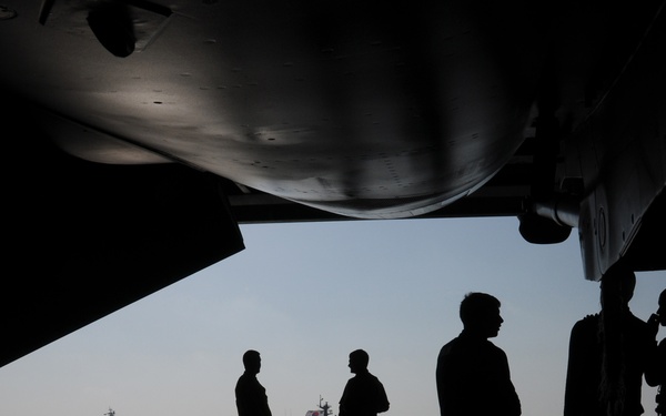 The image shows silhouetted personnel standing on what appears to be the deck or hangar bay of a large naval vessel, likely an aircraft carrier, with a military aircraft visible overhead. In the background, at least two warships are visible on the water, one of which appears to display a South Korean flag. The image is a high-contrast backlit photograph emphasizing silhouettes against a bright ocean horizon.