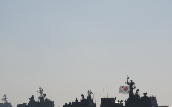 The image shows a formation of multiple naval warships sailing in open waters, silhouetted against a bright, hazy sky. The foremost vessel prominently displays the South Korean national flag (Taegukgi), indicating Republic of Korea Navy participation. The ships appear to be conducting a joint naval exercise or formation sailing.