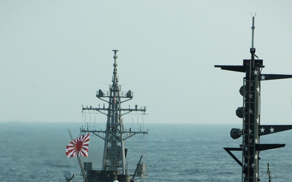 The image shows a Japan Maritime Self-Defense Force (JMSDF) destroyer sailing alongside what appears to be a US Navy aircraft carrier, visible in the foreground. The JMSDF vessel is flying the Rising Sun naval ensign and has Japanese characters on its hull. The scene depicts a joint naval operation or exercise between Japanese and American naval forces at sea.