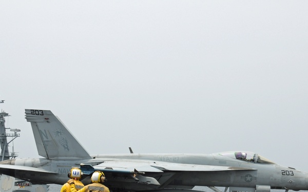 A U.S. Navy F/A-18 Hornet (bureau number 203) is positioned on the flight deck of an aircraft carrier, with two yellow-jacketed deck crew members observing the aircraft. A South Korean naval vessel flying the Taegukgi (South Korean flag) is visible in the background, suggesting a joint naval exercise or operation. The scene depicts standard carrier flight deck operations in overcast maritime conditions.
