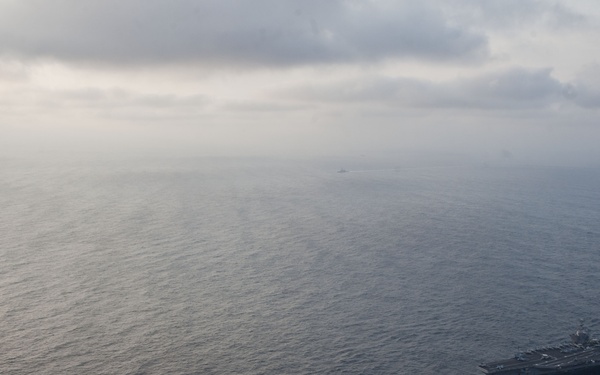 An aerial photograph showing two naval vessels underway in open ocean, with a large aircraft carrier on the right and a smaller warship (likely a cruiser or destroyer) on the left. Both ships are traveling in the same direction, leaving visible wakes in the water. The image appears to be taken from a significant altitude, possibly from a reconnaissance aircraft or helicopter.