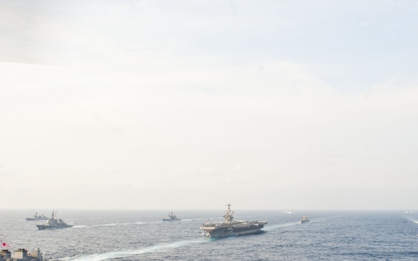 A naval formation of multiple warships is underway at sea, including a large aircraft carrier in the center and several escort vessels. The formation includes at least one Japanese Maritime Self-Defense Force (JMSDF) destroyer, identifiable by the Japanese flag and hull number visible on the nearest vessel. This appears to be a multinational naval exercise or transit operation.