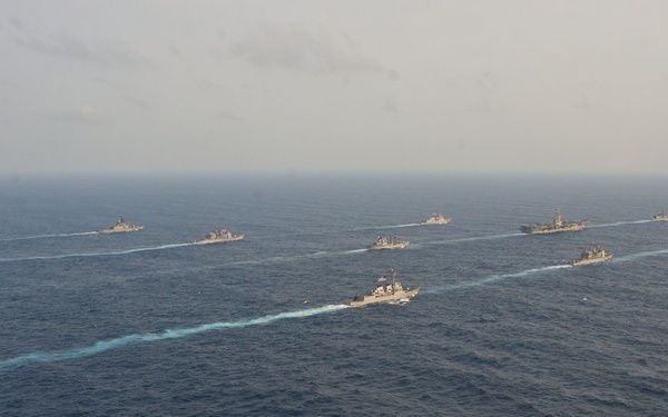 An aerial photograph shows a large naval formation of approximately 10-12 warships sailing in coordinated formation across open ocean. The vessels appear to be traveling in a roughly diagonal line formation, leaving visible white wake trails on the dark blue water. This appears to be a multinational naval exercise or fleet review based on the organized formation pattern.