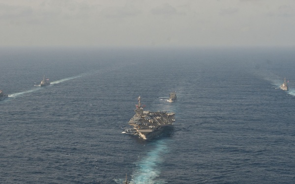 An aerial photograph shows a naval carrier strike group formation underway at sea, with a large aircraft carrier positioned centrally and multiple escort vessels arranged in a protective formation around it. The ships are leaving visible white wakes as they travel through open ocean waters. The formation appears to consist of approximately seven to eight warships of varying sizes.