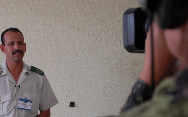 A man in a light-colored military or official uniform with green epaulettes and a name badge is being filmed or photographed against a textured white wall. A second person in camouflage military attire is operating a video camera in the foreground, facing the uniformed subject. The scene appears to be a formal interview or media documentation session.