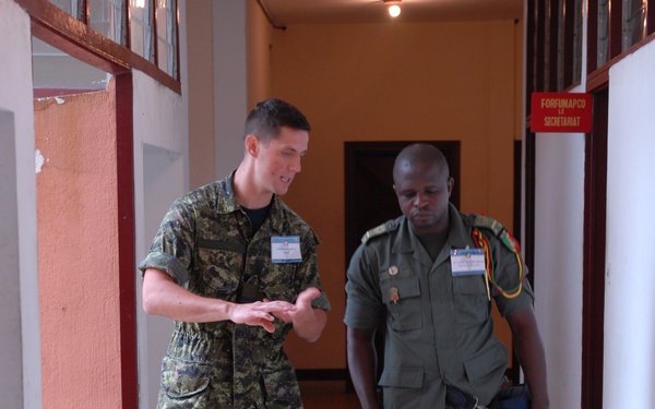 Two military personnel in uniform are walking together down an indoor corridor, appearing to be engaged in conversation. One wears a digital camouflage uniform with tan boots, while the other wears an olive green uniform with black boots and shoulder lanyard. A red sign reading 'FORFUMAPCO SECRETARIAT' is visible on the right wall, suggesting a formal military or peacekeeping meeting venue.