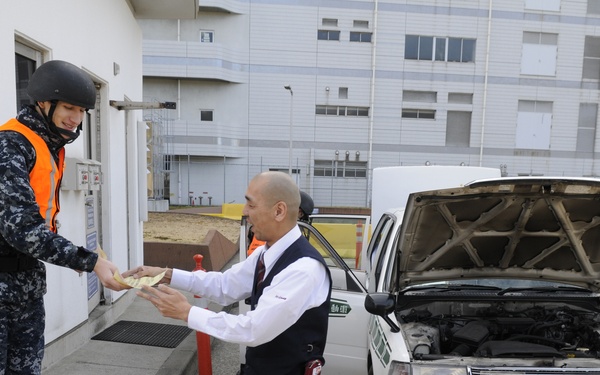 A uniformed security personnel wearing a helmet and orange high-visibility vest is exchanging documents or papers with a civilian driver at what appears to be a security checkpoint. The civilian's vehicle, a Toyota Crown taxi with Japanese license plate 横浜500 あ 98-68, has its hood open, suggesting an inspection is underway. The setting appears to be a controlled entry point, likely at a military or government facility in Japan.