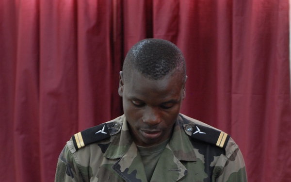 A military officer in camouflage uniform is standing in what appears to be a conference or training room, examining a name badge or credential. He is wearing epaulettes with gold bars indicating officer rank, and a flag emblem is visible on the badge he is holding. The setting includes a laptop, tables, chairs, and red curtains in the background.