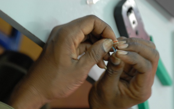 A person wearing military camouflage uniform is working with small components, appearing to manipulate or assemble a small wire or connector with their hands. The close-up shot focuses on the hands and the fine detail work being performed. Various tools and equipment are visible in the blurred background on what appears to be a workbench.
