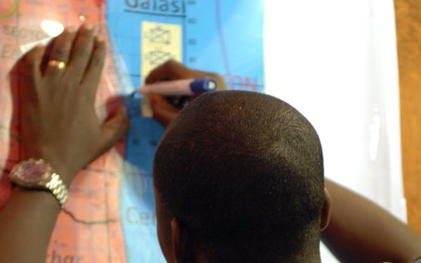 A person wearing woodland camouflage military uniform is working on a large tactical map from behind. A second person's hands are visible on the left side, wearing a watch and ring, also interacting with the map. The map appears to be a military operational or planning map with colored sectors and grid references.