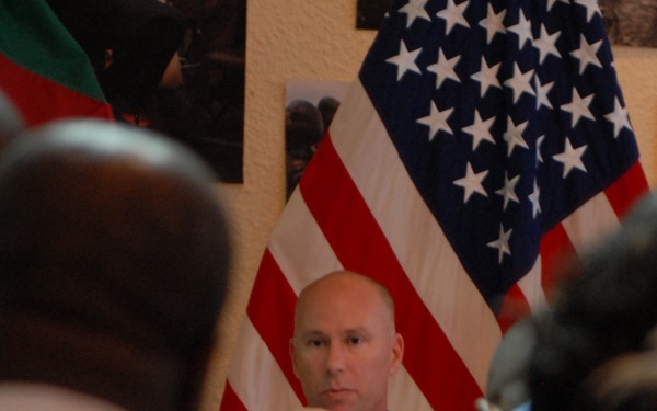 A bald man wearing a military digital camouflage uniform with a name tape reading 'ROBERTS' is seated in front of a large American flag. He appears to be in a meeting or briefing setting, with other individuals visible in the foreground as out-of-focus silhouettes. The setting suggests a military or official gathering.