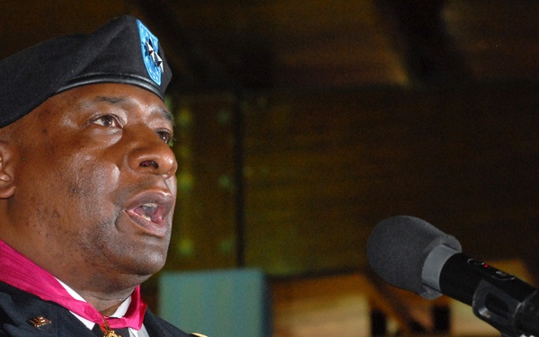 A U.S. Army officer named Hooper, wearing dress blues with multiple medals and decorations including a prominent red/magenta neck ribbon medal, is speaking at a microphone during what appears to be a formal military ceremony or event. He is wearing a black beret with a unit insignia and has a name tag reading 'HOOPER'. The setting appears to be an indoor venue with warm lighting in the background.