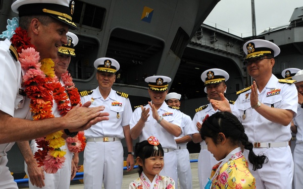 A naval ceremony at a port where a senior officer wearing a floral lei is being greeted by two young girls dressed in traditional Korean hanbok attire. Multiple naval officers in white dress uniforms are applauding in the background near a large naval vessel. The scene appears to be a formal welcome or port visit ceremony.
