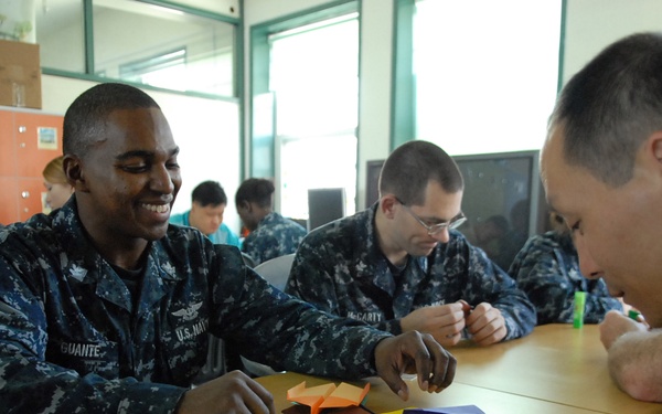 U.S. Navy personnel in digital camouflage uniforms are engaged in an origami activity alongside civilians, likely at a community outreach or cultural exchange event. The setting appears to be a classroom or school environment, with colorful paper folded into various shapes on the table. One sailor, identified by the name tape 'GUANTE,' is smiling broadly while participating in the activity.