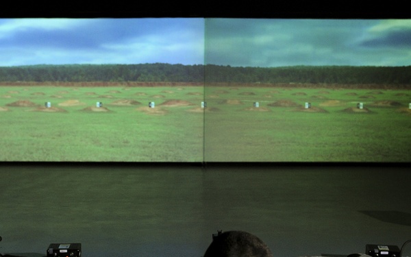 Three military personnel in camouflage uniforms are lying in prone shooting positions facing a large dual-panel projection screen displaying a virtual firing range environment. This appears to be an indoor marksmanship simulation training facility, where soldiers practice shooting skills using simulated weapons and projected targets. The room is darkened to enhance visibility of the projected training scenario.