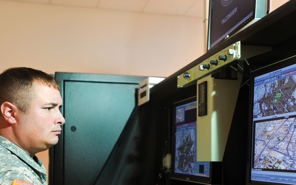 A U.S. Army soldier in ACU (Army Combat Uniform) is operating what appears to be a UAV (Unmanned Aerial Vehicle) ground control station. The workstation features multiple Dell monitors displaying aerial surveillance imagery and mapping software, along with joystick controls. The setting appears to be an indoor military operations or training facility.