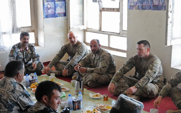 A group of military personnel from what appear to be two different forces are seated on the floor around a low table sharing a meal or meeting. The setting appears to be a simple room with white walls and windows, consistent with a forward operating base or local building. The gathering has the appearance of a joint coordination or liaison meeting over refreshments.