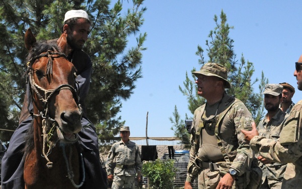 The image shows a group of military personnel in camouflage uniforms interacting with local civilians in an outdoor setting. A local man wearing traditional clothing is mounted on a brown horse, appearing to demonstrate or present the animal to the soldiers. The scene appears to take place in a grassy area surrounded by pine trees under clear blue skies.